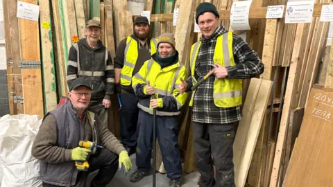 Eimert is pictured with their volunteers. Most are wearing high-viz bibs and caps and work wear as they stand in front of many wooden panels, smiling with some giving the thumbs up.