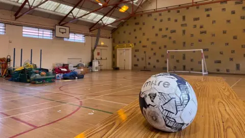 A black and white football on a wooden table in an abandoned sports hall. PE kit can be seen on the wooden floor in the background. 