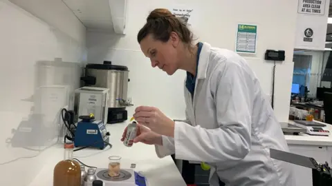 Jess Warren/BBC A woman in a lab coat ​uses a pipette to drop a liquid into ‌a beaker.She stands in a research and growth lab.