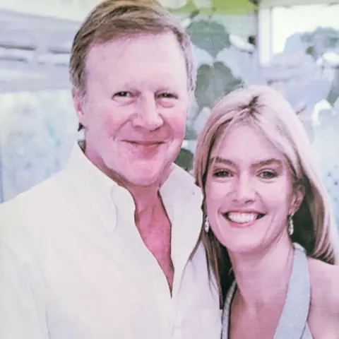 The Good Grief Trust Graham Theakston and Linda Magistris pose together for the camera. Graham is taller than Linda and has short brown hair and is wearing a white open collar shirt. Linda has long straight blonde hair and is wearing a light blue halter neck top.
