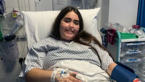 Lucy Harman A young woman with long dark hair wearing a hospital gown lying in a hospital bed, with an intravenous canula in her hand and a nasal canula in her nose