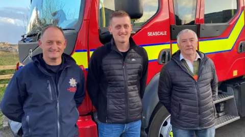 From left Terry Steele, his son George, and his father Chris. Terry is a middle-aged man who has thinning grey hair and is smiling, and wearing a fleece with the Cumbria Fire and Rescue Service crest on it. George is younger and the tallest of the three, has short fair hair and is wearing a puffa-jacket style fleece. Chris has short grey hair and is also wearing a black puffa jacket. They are all standing in front of a fire engine.
