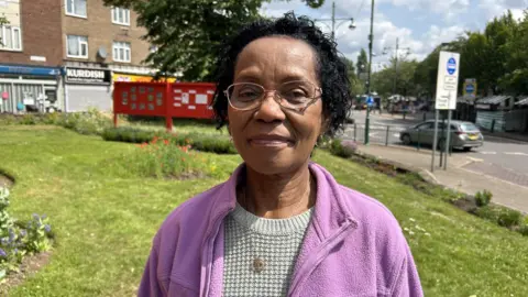Norma Thompson in Wednesfield town centre. We can see some shops and cars behind her. She has short black hair and she is wearing a purple fleece and a grey jumper. She has a necklace on and is wearing glasses.