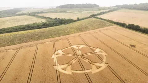 Lewis Johnstone Photography An aerial view of a crop circle resembling a five-spoked wheel with a rural landscape in the background