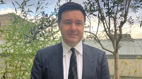 Dave Malone is standing outside with leaves, a tree and flowers behind him. He's wearing a grey suit with a white shirt. 