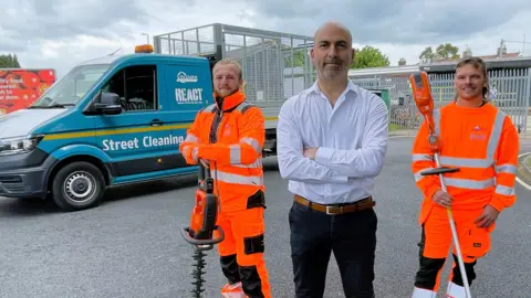 There are three men standing in front of a van which says React - street cleaning on it. Two men have high visibility suits in orange on and have litter picking and rubbish clearing tools in their hands. The third man is wearing a white shirt and black trousers and has his arms folded. 