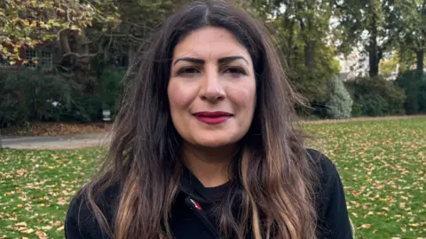 A woman with long dark hair, wearing red lipstick and a black coat stands in front of a green space with trees and shrubs behind her