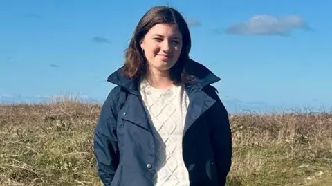 Youth Commission Guernsey A picture of Eva stood in a field. She is wearing a navy coat and is surrounded by grass.