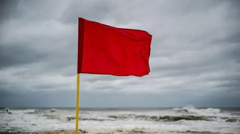 Getty Stock image on red flag on yellow flagpole in front of ocean. Sky is filled with clouds. 