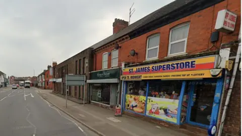 Google An urban street of brick two-storey shop units with a pavement and road in front. The one closest to the camera has a panel above the door saying "ST JAMES SUPERSTORE".