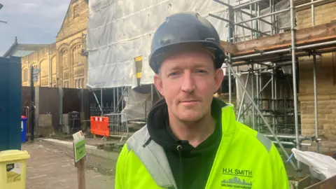 Photograph of Mark Taylor from H.H. Smith & Sons. He is pictured in a hard hat and a high-vis jacket on a building site.