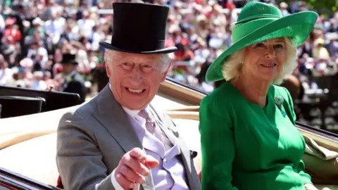 Reuters The King and Queen arrive at Ascot in the first carriage of the Royal Procession. The King is wearing a black top hat and pointing with his right hand. The Queen is wearing a green hat, matching her outfit.