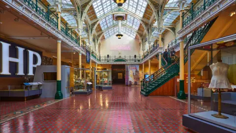 David Rowan/Birmingham Museums Trust Glass-roofed building with red and blue floor and objects displayed in cabinets. On the right is a green staircase that leads to the second floor.