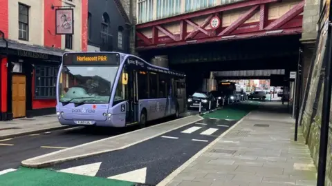 A purple-coloured bus, ahead of other cars, drives past a floating bus stop. There is a small paved section between the main road and the cycle lane, with a pavement on the right hand side. It is situated next to a pub and underneath a red-coloured railway bridge.