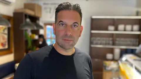 Deli owner John Hyman inside shop in West Bridgford, Nottinghamshire, wearing a dark t-shirt and looking at the camera.