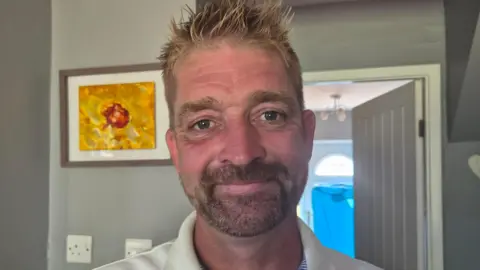 An older man with blonde hair and a dark beard smiling at the camera