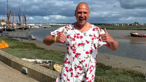 Matt Mackay/BBC A man smiling, dressed in a white dress with red flowers, with his thumbs up