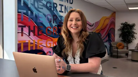 Mel Brooker sat behind a black desk with a rose gold Apple laptop on. She is smiling at the camera and has her hands clasped together in front of her. She has long, curled, ginger hair and is wearing a black, short-sleeved blouse with white lace featured on. She is also wearing a watch and her nails are painted blue. On the wall behind her is a large, colourful graphic with "THE GRIND" written multiple times.