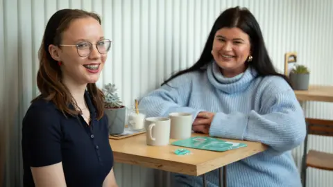 BBC Kitty and Jessie sat at a table in a coffee shop. Kitty has long dark hair and is wearing a black headband. She has glasses and braces and is wearing a navy t-shirt. Jessie has long black hair and gold hoop earrings, and is wearing a light blue knitted roll neck jumper. 