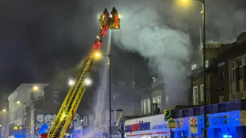 PA Media A cherry picker being used by London Fire Brigade containing two firefighters in the top platform spraying water on to the building below. There is a row of shops and the air is smoky. It is also dark as it is early morning.