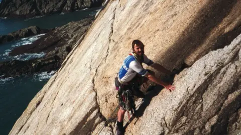 Mike Wigney A picture of Mike with dark hair and a beard climbing an almost vertical smooth slope with the sea below. He is wearing climbing gear.