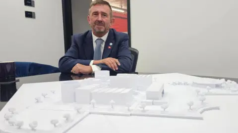 Michael Keohan/BBC A man in a blue suit and white shirt and light blue tie. He is sitting in front of a 3D model of a proposed hydrogen plant. 