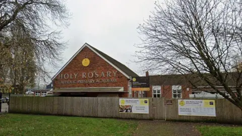 A red brick school building with a pointed roof behind a wooden fence. There is a yellow circular sign on a wall of the building, with a large sign in bronze letters that reads "Holy Rosary Catholic Primary Academy"