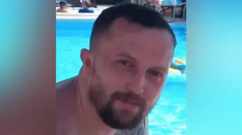 A man is sitting by the pool and looking into the camera. There's a float in the water behind him.