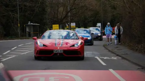 A red super car in front of two other cars driving along a street close to a family walking on a pavement. 