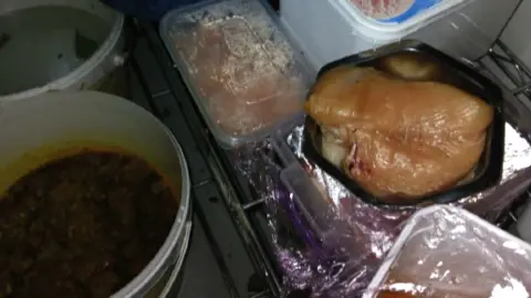 Cardiff Council A raw, dated chicken breast is uncovered in a walk-in fridge sat on a container on a shelf. Besides it are other foods which are covered in containers. On the floor is a large tub of an indescribable brown food and liquid which is uncovered. 
