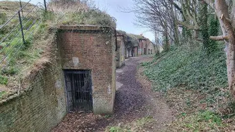 Old brick buildings build into a hillside on the left with a winding mud path to its right, which is lined by trees and ivy
