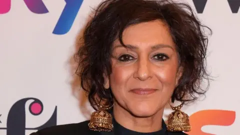 PA Media Meera Syal is looking at the camera, in front of a wall with brand names on it. She has her hair up and is wearing large gold earrings,