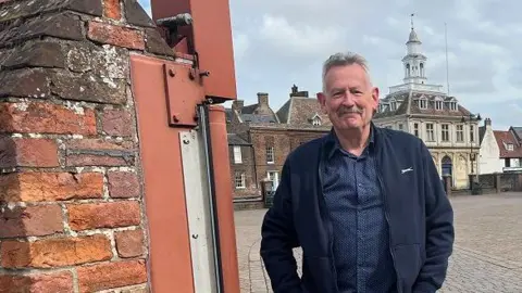 BBC Heritage Open Day organiser Kim Leonard stands by a brick wall in front of King's Lynn's historic Custom House. He wears blue denim jeans, a blue shirt and blue fleece. He has grey hair and moustache. 