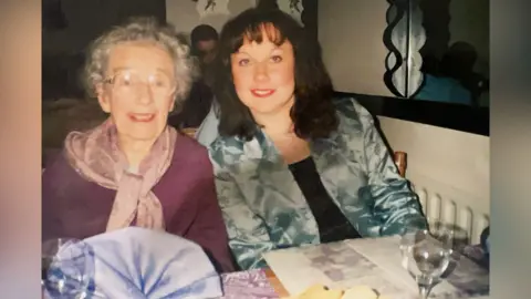 The Fryd Family Elly Downes with her grandmother Judy Fryd sit at a restaurant table side by side. there are napkins, menus and a wine glasses on the table. A man can be seen sitting on a table behind them. An eldely Fryd and her grand daughter are smiling at the camera. 