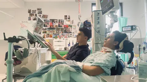 Jelenyela Pena Seb in his hospital bed, with lots of equiptment atached to him. His brother is sat next to him and scrolling on a laptop propped up in front of Seb. In the background is a wall covered with images of Seb with his friends and family.