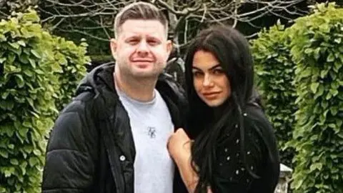 Sussex Police Josh and Chloe Bashford embrace looking towards the camera in a garden. On the left Josh has short light brown hair and a stubble beard. He wears a grey t-shirt and a black puffer coat. On the right Chloe has long black hair and wears a black jumper with diamante on the sleeve. 