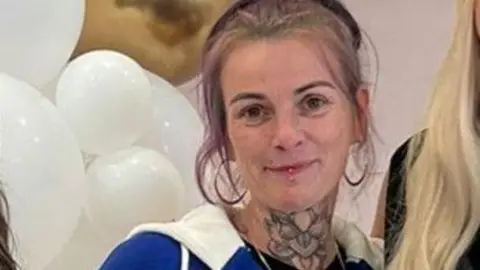 A zoomed in shot of a woman who has fair hair with light purple highlights. She is wearing large hoop earrings and a blue and white tracksuit top. The long blonde hair of someone next to her can be seen as well as some white balloons behind her. 