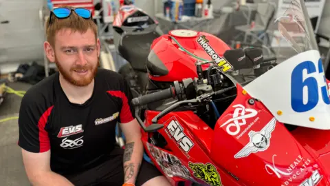 Grant Thomson crouches next to a red motorbike in a workshop. He has ginger hair and a ginger beard. He wears sunglasses on top of his head and a black T-shirt with white logos on the chest. 