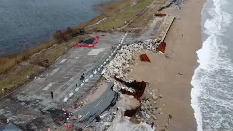 An aerial view of the road at Torcross which has been destroyed by storms. Twisted sheets of metal are visible and large amounts of rocks which are now where the road previously was. The sea is on the right and the lake on the left of the road.