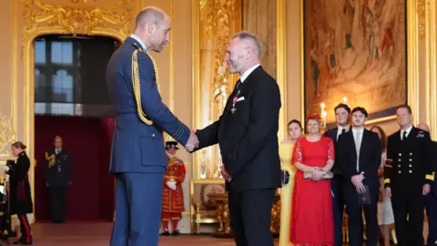 PA The Prince of Wales and Gary McKee shake hands as McKee is awarded an MBE at a ceremony at Windsor Castle. They are both formally dressed. A group of guests is standing in front of a large painting in the decorated room.