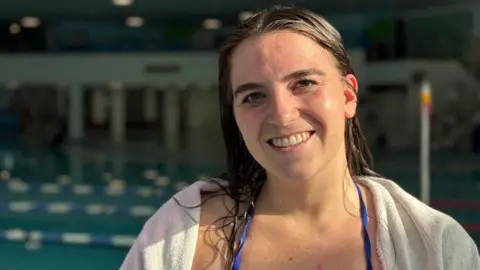 Orla Rabey is smiling as she stands by the pool. She has long brown wet hair and is wrapped in a white towel.