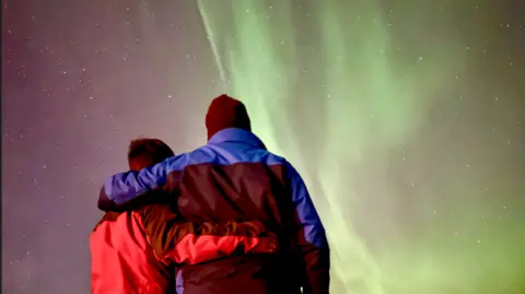 Zak Nelson Two figures with backs to camera and arms around each other, one wearing a red waterproof jacket and other a black and blue waterproof jacket and black woolly hat, with the aurora of green lights in the sky in the background