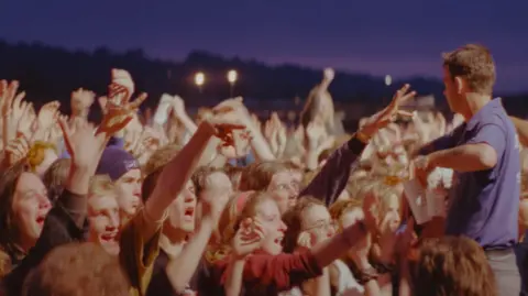 Mensch Films/Robin Mahoney A close up of a group of festivalgoers at night, lit up from the stage, all with their hands in the air, and some are reaching towards drinks, which a stage-hand is passing to them