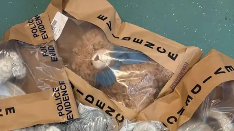 Nottinghamshire Police A close up of a police evidence bag with a brown teddy bear inside. The bear has a blue bow and can be seen through a plastic window in the brown bag. The brown bag has a large label down the side which reads 'EVIDENCE'
