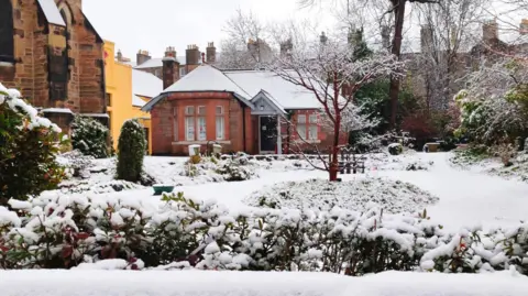 Aditi Mazumder A house covered in snow. The house is made of red brick and has a large ground floor bay window. The roof is covered with snow, as is the garden in front of it. Some greenery from bushes can be seen in the foreground. A reddish-brown tree, which is bare, has snow on its branches. On the left of the image, the corner of a church, made of light and dark stone, can be seen.