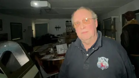 Paul Fowler is wearing a black shirt and glasses. He has short grey hair and is standing in front of a aircraft memorabilia.