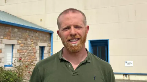 A man wearing an olive coloured polo shirt smiling standing in front of a white and stone building.