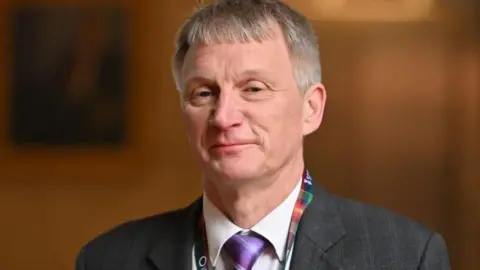 Getty Images Ivan McKee caput  and shoulders wearing a purple necktie  and a lanyard