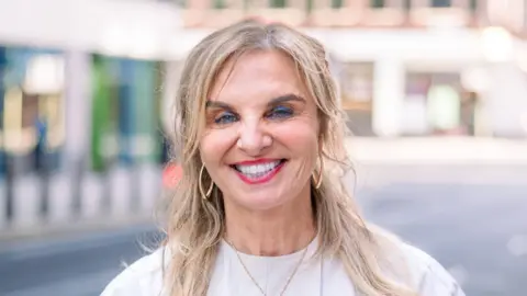 Andrea Minichiello Williams A woman wearing blue eye shadow smiles at the camera. Her shoulder-length hair is worn down and she has gold hoop earrings in. The background is out-of-focus but sunny London street.
