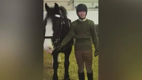 PA Media Jack holds the reigns of a black horse with a white patch on its face, and smiles at the camera. He is wearing a riding helmet, a military green knitted jumper with a belt and brown riding trousers and boots. Hay can be seen on the floor, with a stable behind him. 